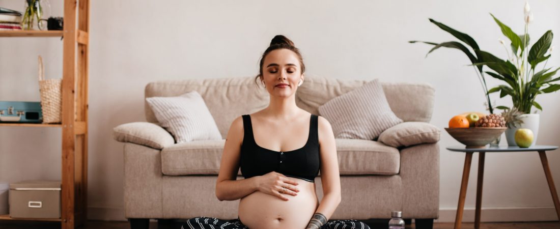 Cheerful brunette pregnant woman enjoys music in white wireless headphones and gently touches belly. Cheerful tanned girl in black pants and top meditating on purple carpet