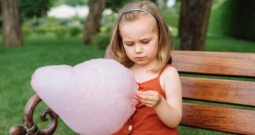 Cute toddler girl eating big pink cotton candy in the park on a summer sunny day.