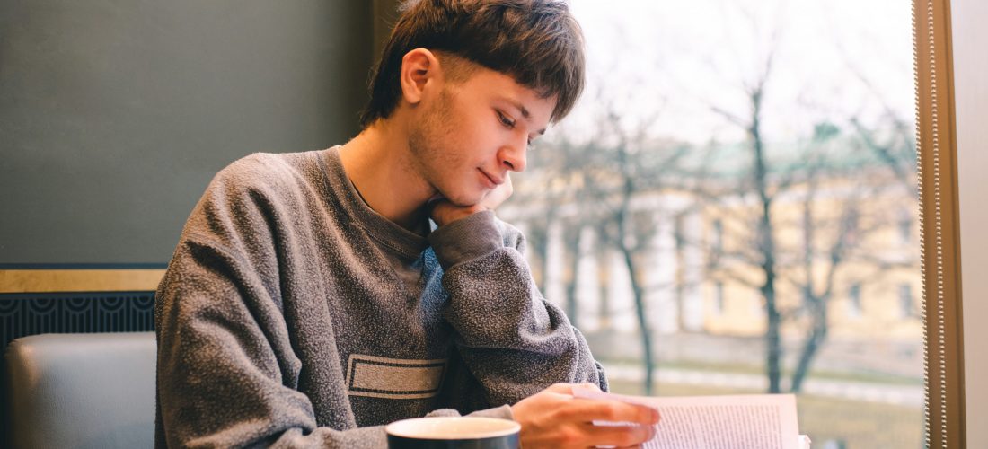 Teenage blond boy 18-19 year old reading book and drinking coffee in cafe alone.