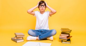 Portrait of attractive young little small cute shocked school boy with books, wearing glasses, palms to head, doing home work, over yellow background, isolated