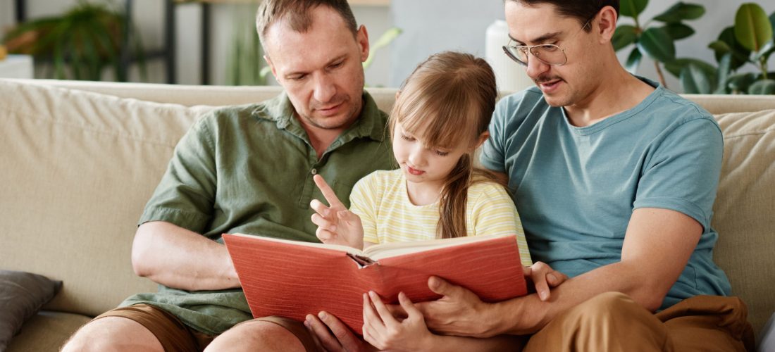 Gay parents sitting on sofa and reading a book to their little daughter in the room